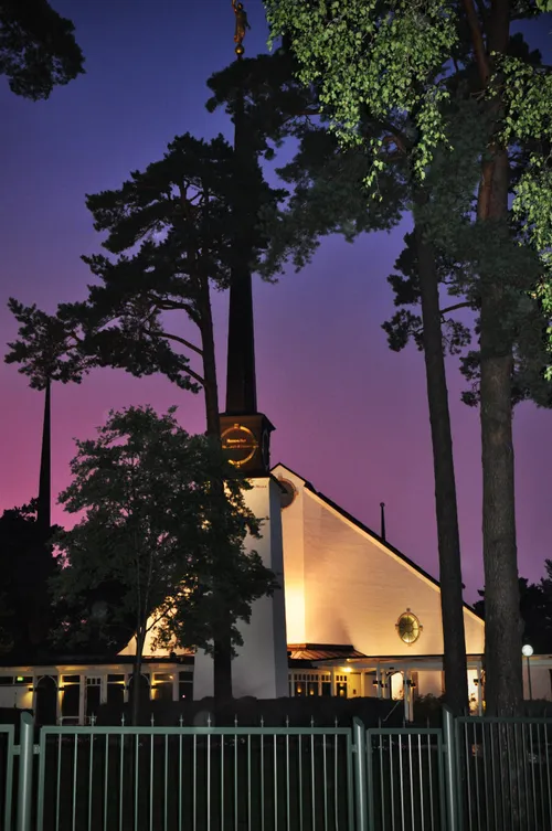 The grounds of the Stockholm Sweden Temple, including the fence and trees, with the temple lit up in the background.