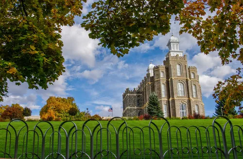 The Logan Utah Temple on the hill, with the temple’s silver fence in the foreground and green tree branches overhead.