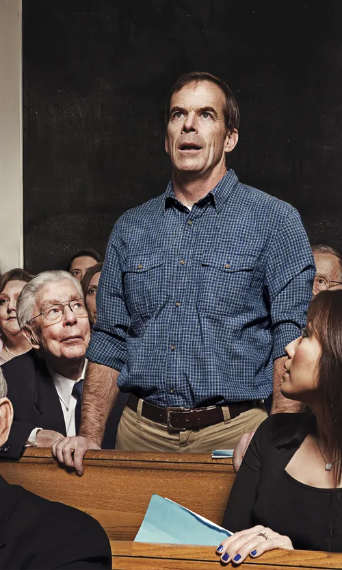 A middle aged man stands and speaks from the pews. Others are watching him.