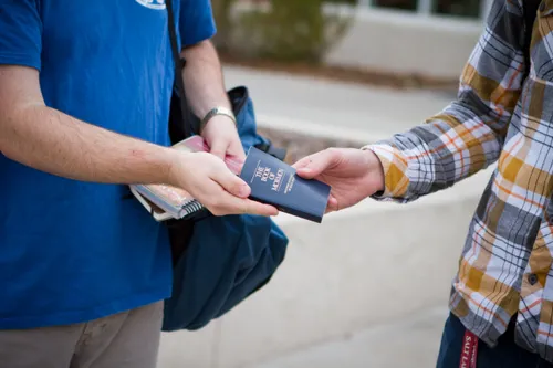 The hands of a young man giving a paperback Book of Mormon to another young man.