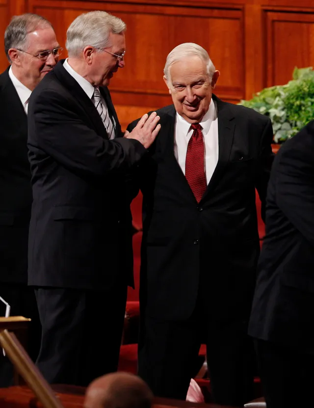 Elder D. Todd Christofferson places his hand on Elder Richard G. Scott's shoulder as they stand and acknowledge people in the audience. Elder Scott is smiling.