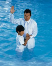 A young boy in a white jumpsuit standing in a large body of blue water to be baptized by a man who is raising his arm to the square.