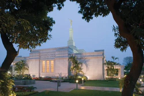 A view of the Asunción Paraguay Temple in the evening, seen between two trees, with the words “Santidad al Señor: La Casa del Señor” above the door.