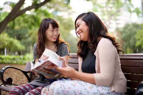 Two young women sit on a wooden bench together outside near trees and read from the Ensign.