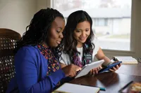 Two sister missionaries sit indoors and read together from tablets.