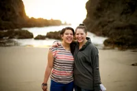 Two women embrace each other on a beach next to cliff side
