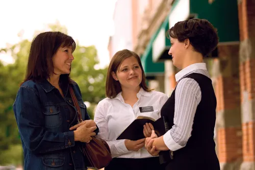 Two sister missionaries holding their scriptures and speaking with a woman wearing a denim jacket in an outdoor setting.