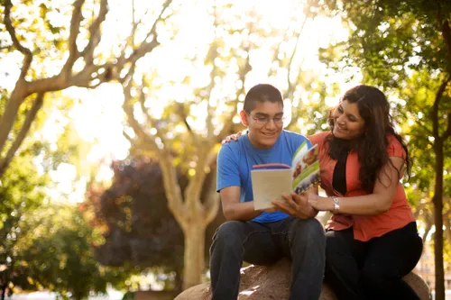 A mother sits next to her son on a rock outside and reads a FamilySearch pamphlet with him.