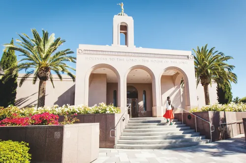 A woman with brown hair, a white shirt, orange skirt, and blue high heels walking up the stairs to a temple past flowers and palm trees.