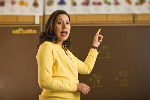 A female teacher with brown hair and a yellow sweater stands at the front of a classroom and points to the blackboard behind her.