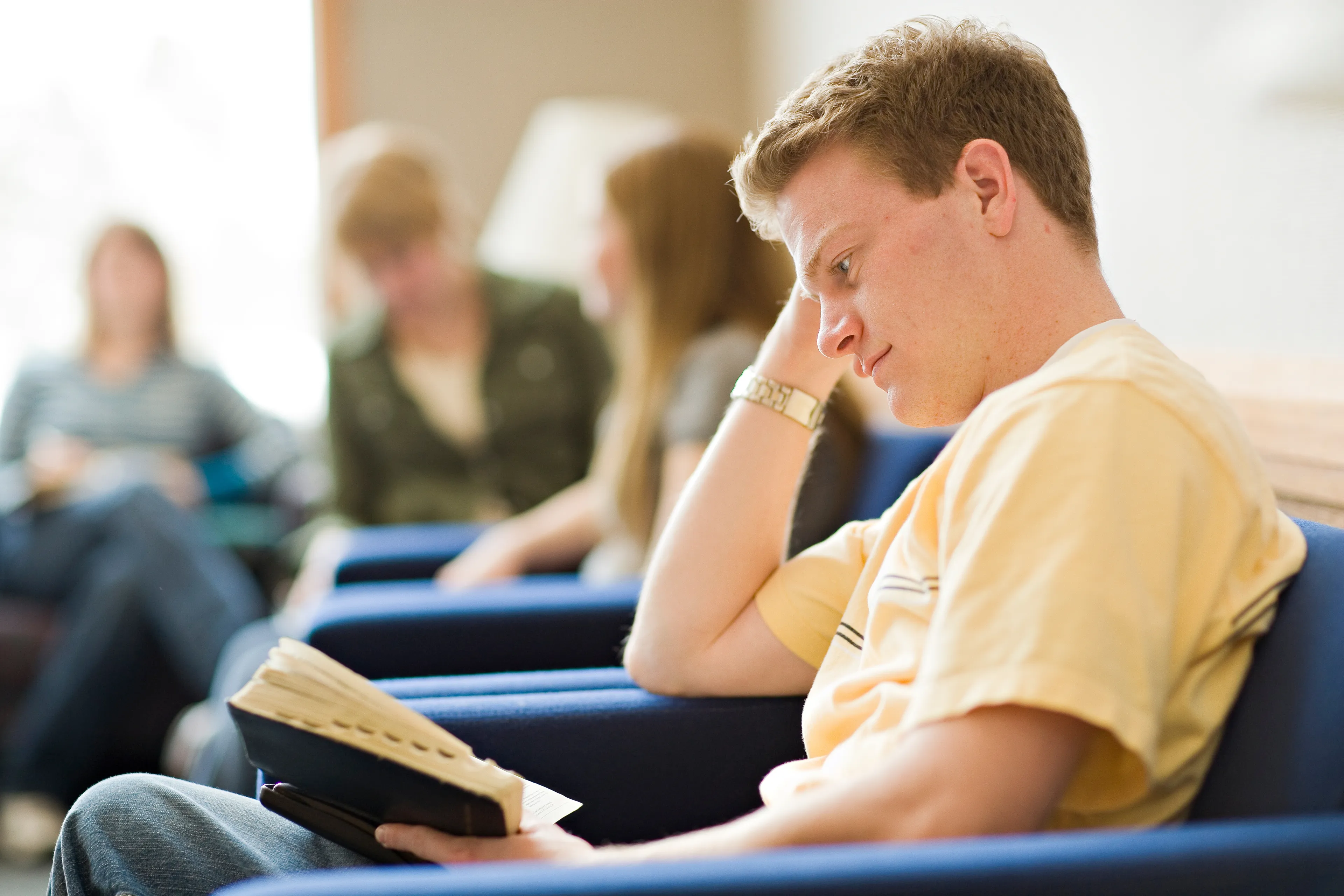A young man sits in a chair and reads from his scriptures.