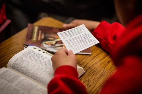 A student’s hand is seen holding a piece of paper with a quote typed on it, with his scriptures laid open on his desk next to a workbook.