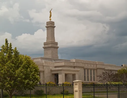 A front view of the Monticello Utah Temple entrance, fences, and green grass and trees, with a stormy sky.