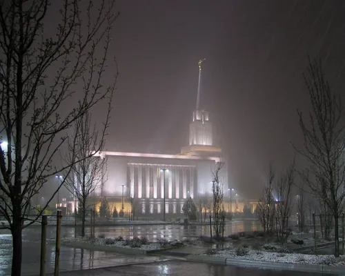 A side view of the Oquirrh Mountain Utah Temple on a snowy evening, with bare trees in the parking lot medians.