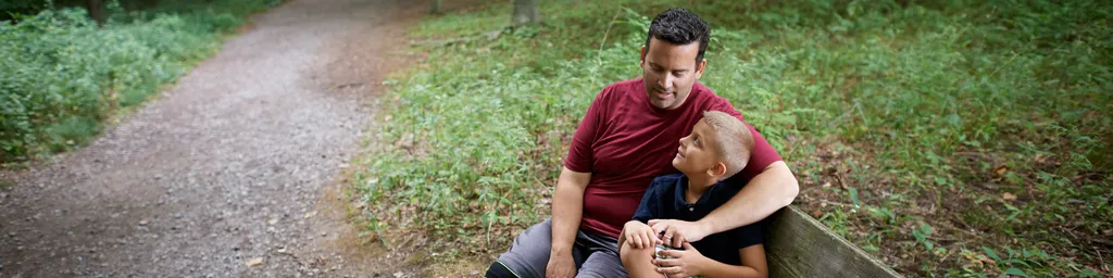 Various people sit on a bench off pathways around the Scared Grove. There are lots of trees and dirt pathways surrounding.