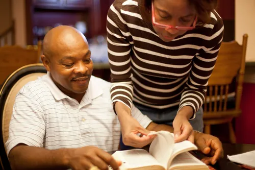 A woman with a striped shirt and glasses standing by a man in a chair and helping him study the scriptures at a table.