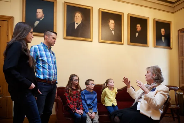 Various families are inside the Beehive House (home of Brigham Young) in Salt Lake City, Utah. They all interact with the exhibits in the museum. There is a docent/missionary talking with them.