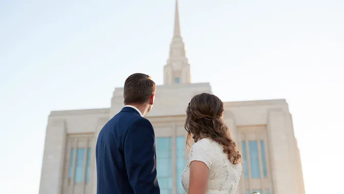 Young couple stands outside the Ogden Utah Temple. They are looking out at the temple in the background with the Angel Moroni being almost top center of the image. He is wearing a dark blue suit with white collared shirt. She is in a white dress. It is late afternoon/early evening.