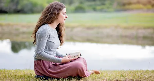 Young adult woman ponders scriptures in a pastoral setting near a lake.