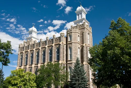 A side view of the Logan Utah Temple, with a partly cloudy sky in the background and several trees growing near the temple walls.