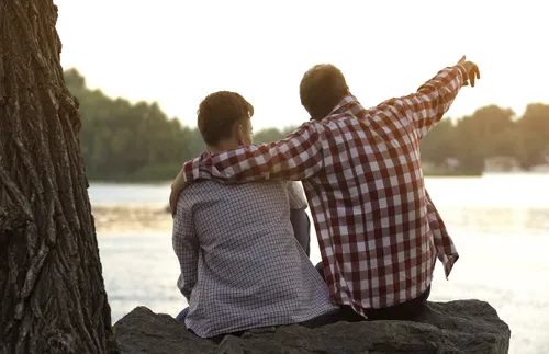 father and son sitting on riverbank, with dad pointing at distance