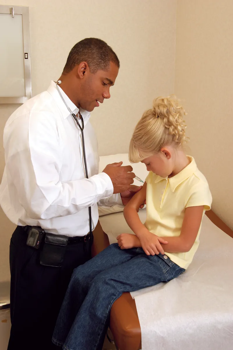 A doctor standing and giving an injection to a young girl sitting on a bed.