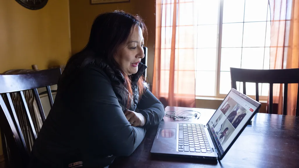 A woman sits at her kitchen table and works on her laptop. She appears to be attending a virtual meeting.