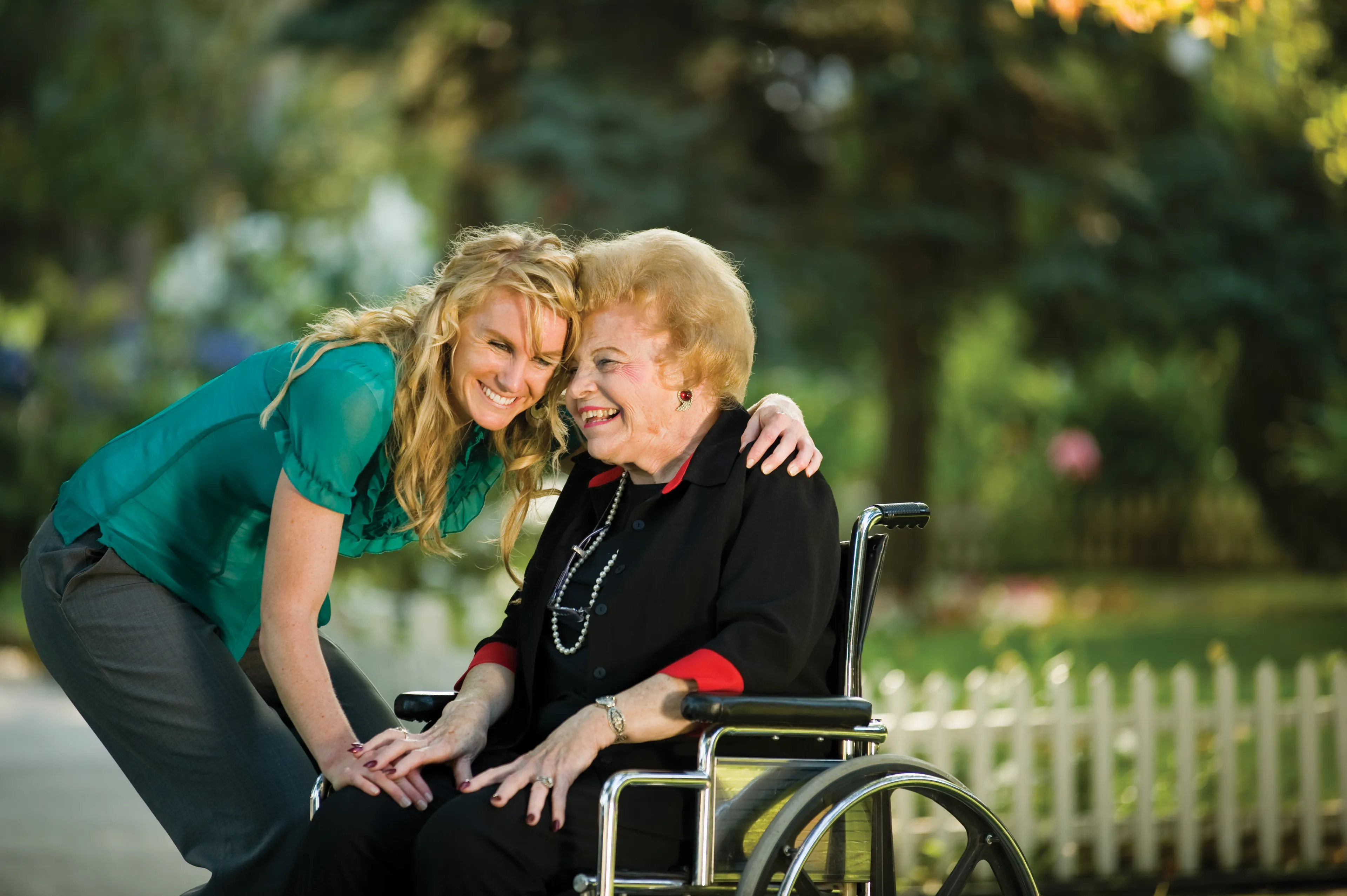 A woman bends down and hugs an elderly woman in a wheelchair.