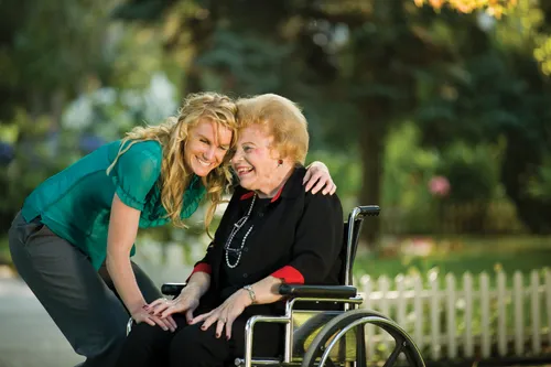 A blonde woman in a blue shirt and gray pants smiles while hugging an elderly woman in a black shirt and pants, who is sitting in a wheelchair outside.