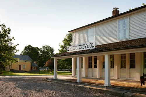 White nineteenth-century building with a broad covered walkway in the foreground. Yellow clapboard house with a red door in the distance.