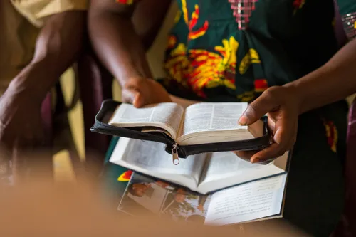 A woman wearing colorful clothing and holding open a set of scriptures in a zipper-closure case. Another book of scripture is on her lap.