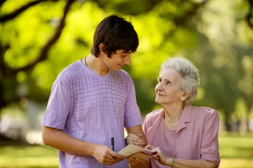A young man holding a notebook smiles while walking arm in arm with an elderly woman outside.