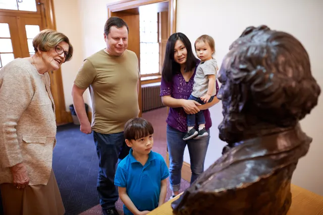 Various families are inside the Beehive House (home of Brigham Young) in Salt Lake City, Utah. They all interact with the exhibits in the museum. There is a docent/missionary talking with them.