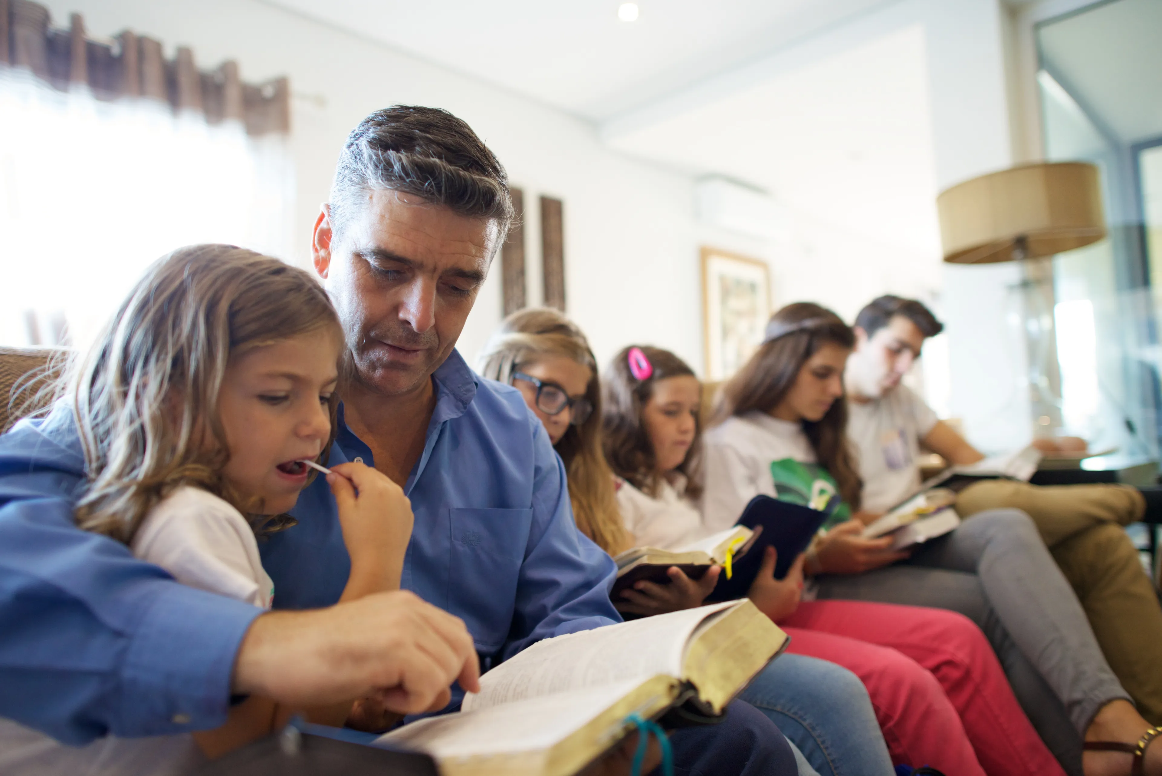 A father sits with his children in their living room to read from the scriptures together.
