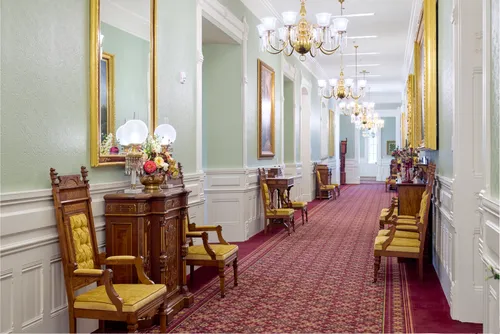 Interior image of the Manti Utah Temple. The image featuring architectural details. This image focuses on a hallway. We see the red and gold carpet as well as the wood chairs with gold accents.