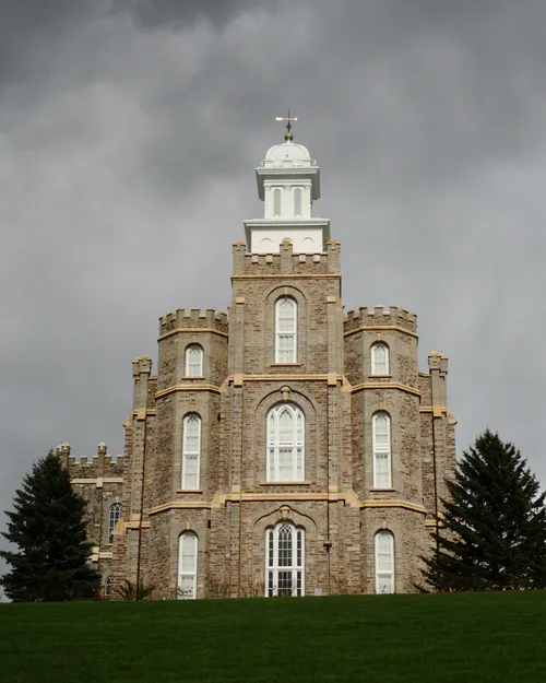 The front of the Logan Utah Temple from the bottom of the hill on a stormy day, with two pine trees on either side of the temple.