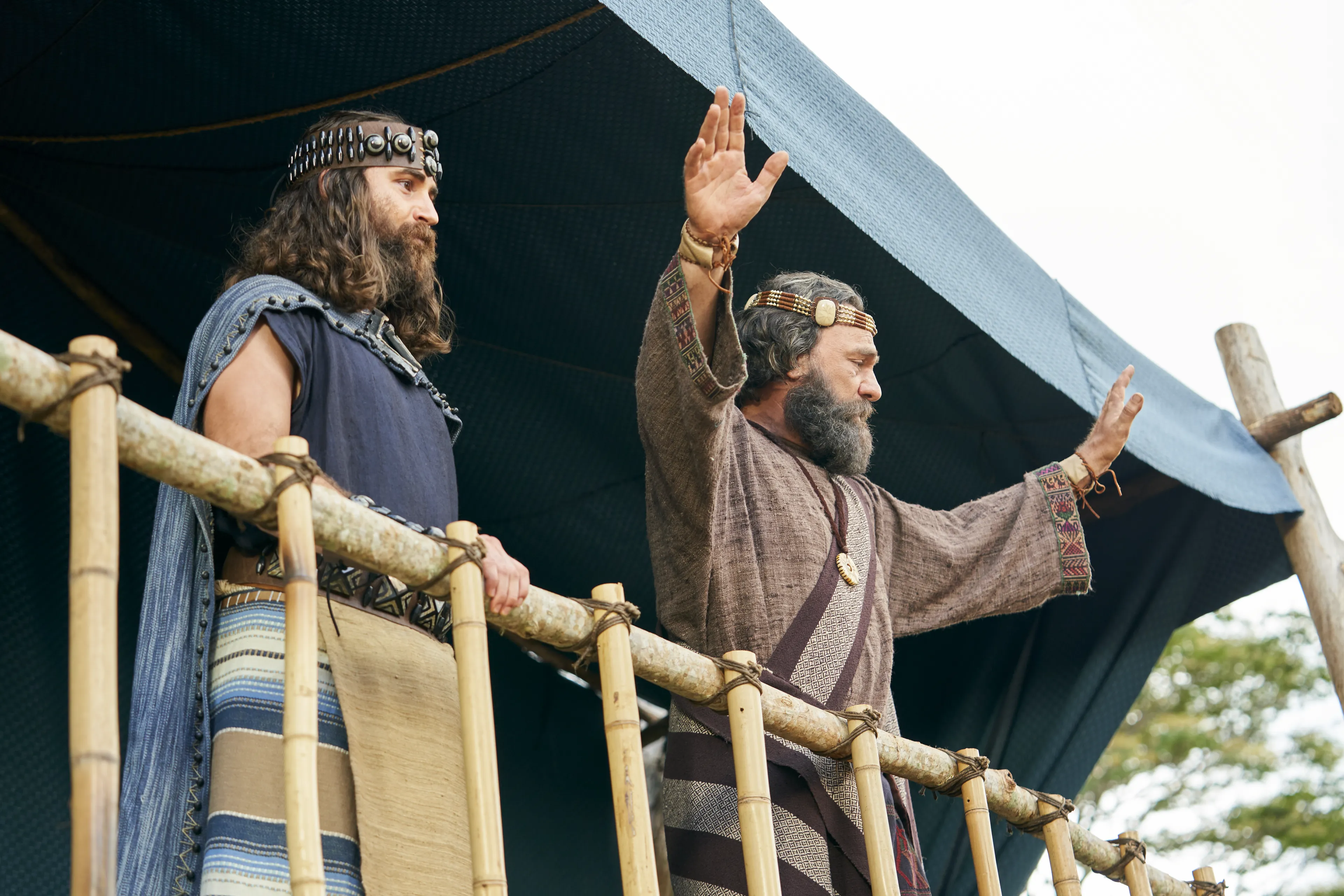 King Benjamin stands on a tower speaking to the Nephites and Mosiah watches in the Land of Zarahemla.