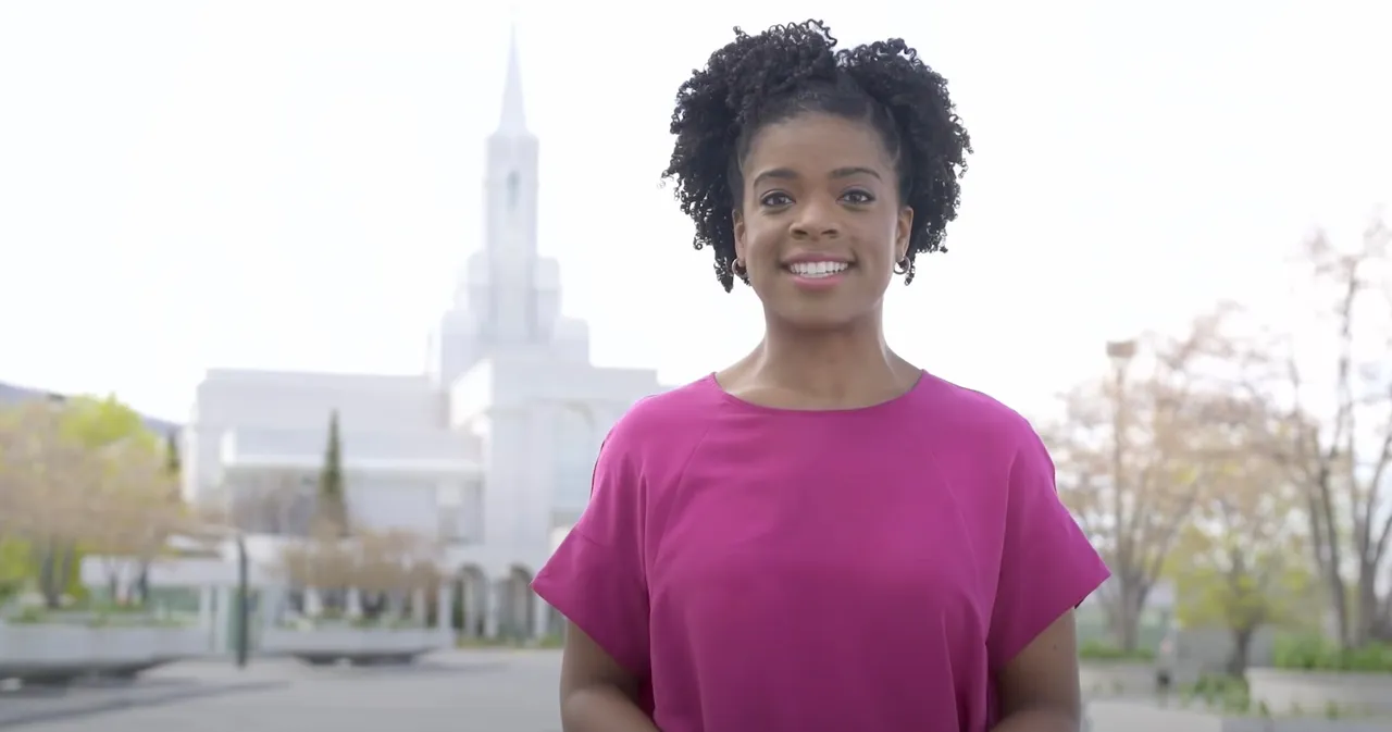 A woman stands smiling in front of temple during spring to explain why baptisms for the dead are preformed in temples