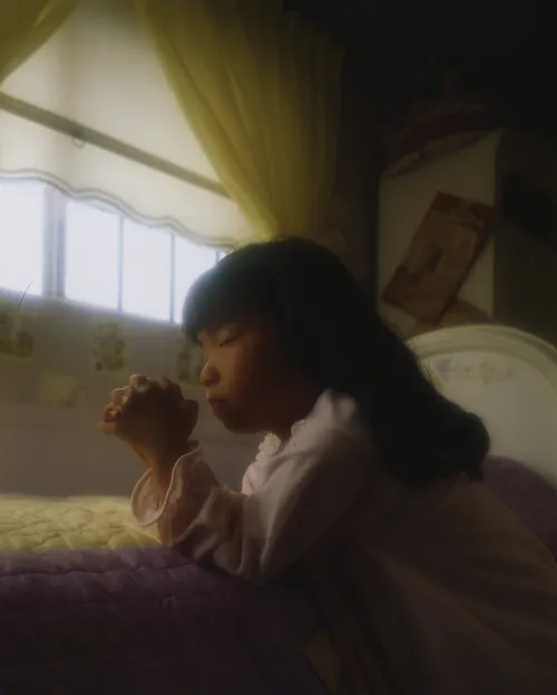 A young girl with black hair kneels at the side of her bed in the morning with her hands clasped and eyes closed in prayer.