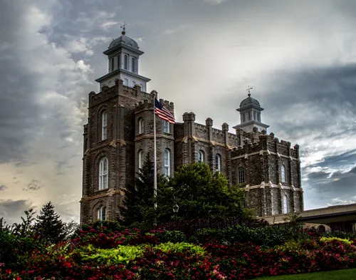 A side view of the Logan Utah Temple, with a dark, cloudy sky above and large green and red bushes in the foreground.
