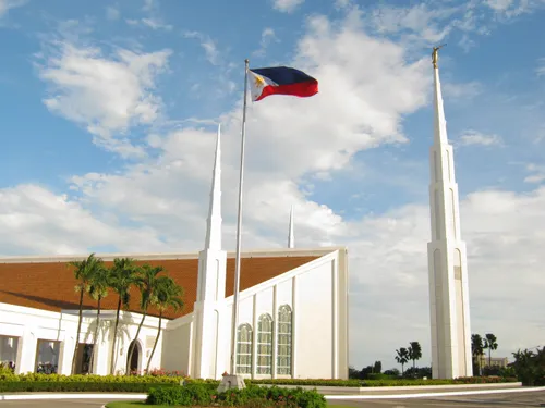 A view of the entrance and grounds to the Manila Philippines Temple, with a separated spire tower to the right and the Philippines flag blowing in the wind on a sunny day.