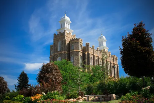 The Logan Utah Temple and grounds on a sunny day, with a large red tree on the right side and a blue sky overhead.