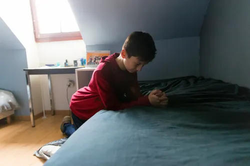a young man kneels at his bedside, head bowed in prayer
