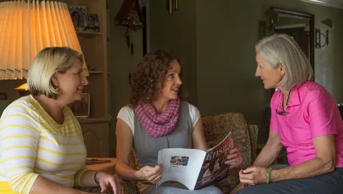 Two ministering sisters sitting on a couch, with one reading from the Ensign to another woman in a pink shirt.