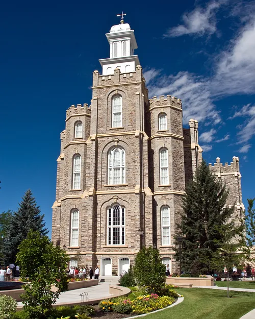 The front of the Logan Utah Temple on a sunny day, with parts of the grounds and pine trees in the foreground.