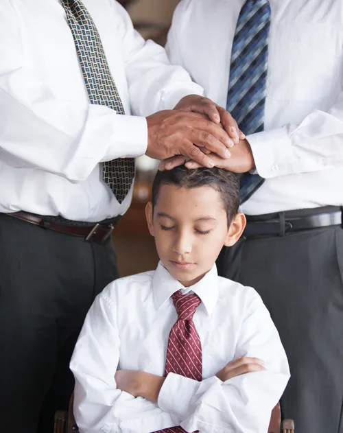 photo of boy with closed eyes and folded arms and two men placing their hands on his head