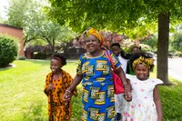 A family walks together outside an LDS Chapel. A mother, brother, and sisters all laugh and walk together, talking. They are all in what appears to be traditional African clothing. Everyone seems happy. The church building is brick with white stone accents. There is a sign on the side of the building which is mounted to the brick.   This is a part of a 2017 signage project showing different kinds of church signs