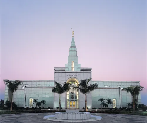 The Campinas Brazil Temple lit up in the early evening, with palm trees near the entrance.