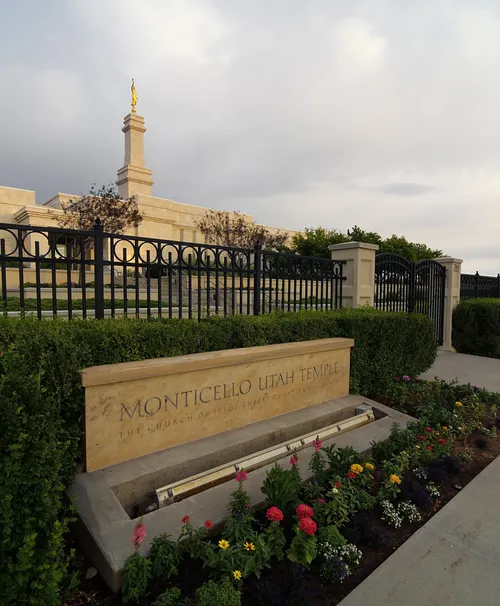 The name sign of the Monticello Utah Temple, with flowers and plants in front of the temple fence.