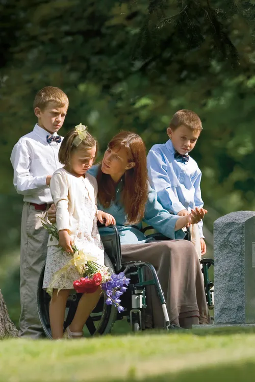 A mother in a wheelchair is surrounded by three young children.  They are in a cemetery in front of a tombstone.